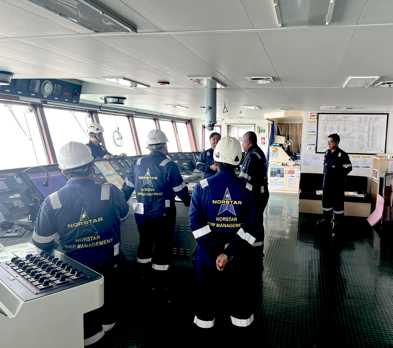 Norstar crew members in navy boiler suits and white safety helmets take part in a seafarer compliance training briefing on a vessel's bridge, gathered around the navigation console with one officer addressing the group.